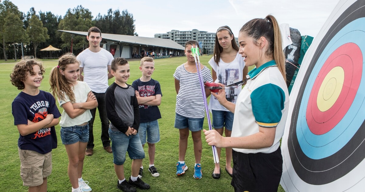 A group of children stands on a grassy field listening to an instructor holding arrows beside an archery target.