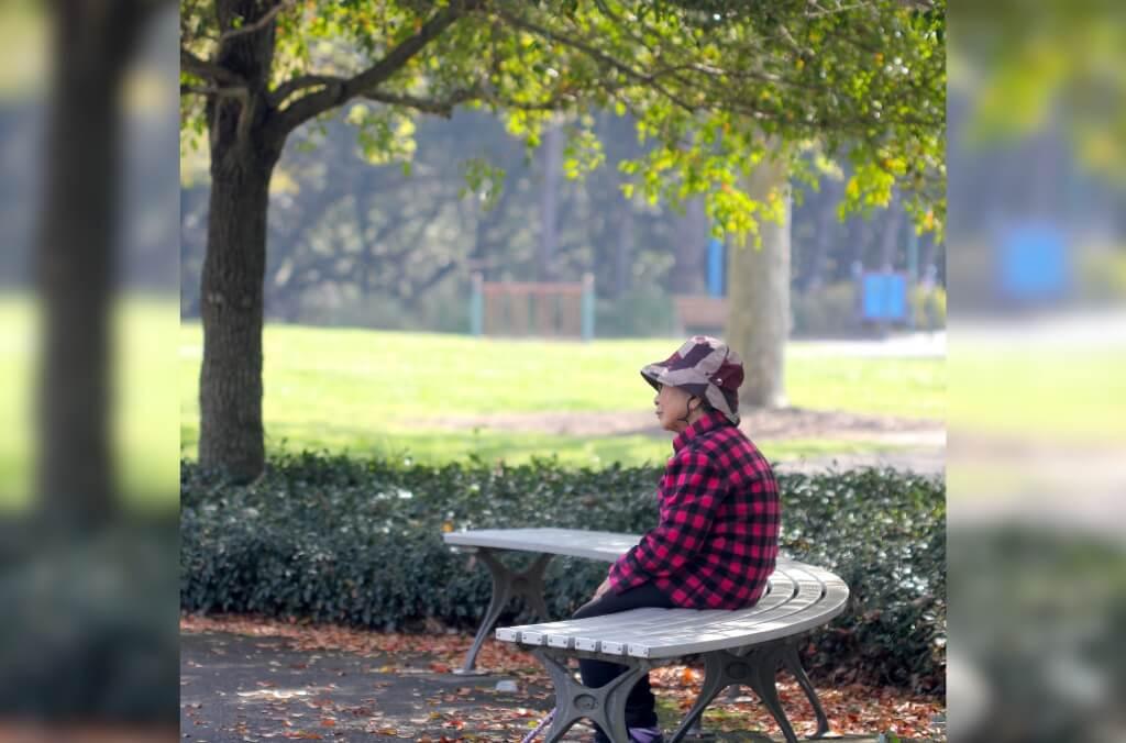 Person in a red and black checkered shirt sitting on a bench in a park, with trees, greenery, and park equipment in the background.