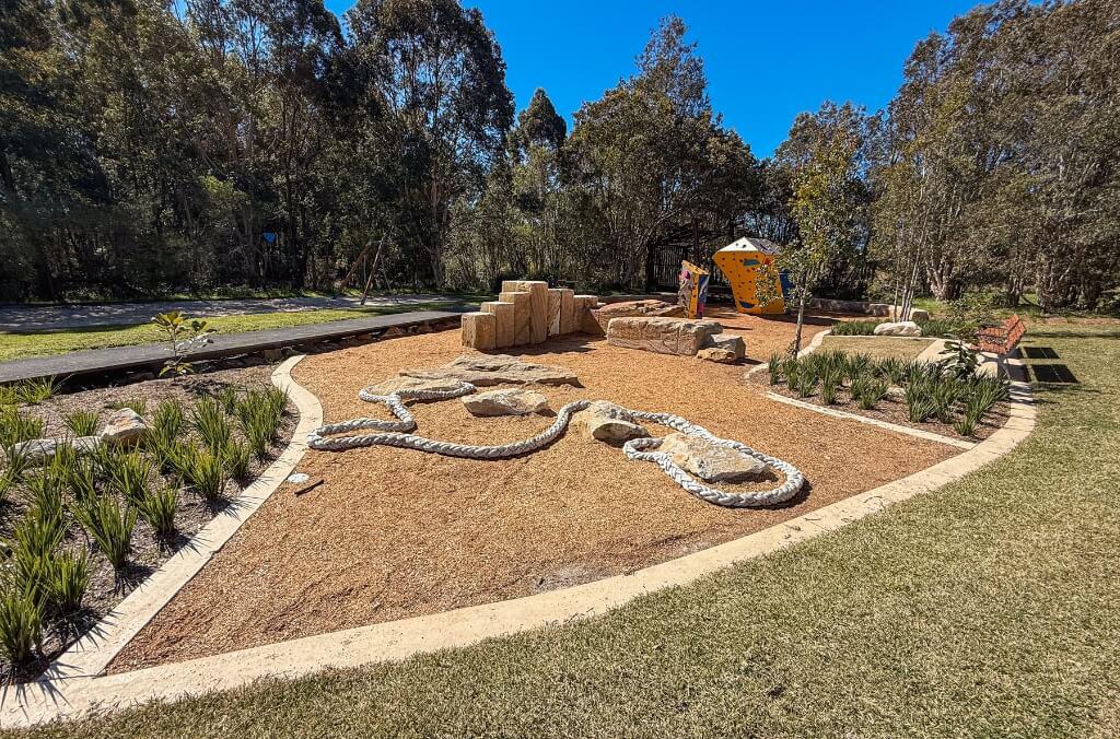 Outdoor playground surrounded by trees, featuring a large rope and rock climbing structure on mulch ground, stone blocks in the background, a yellow climbing wall with colorful holds, and a nearby bench, bordered by grass and landscaped plants.